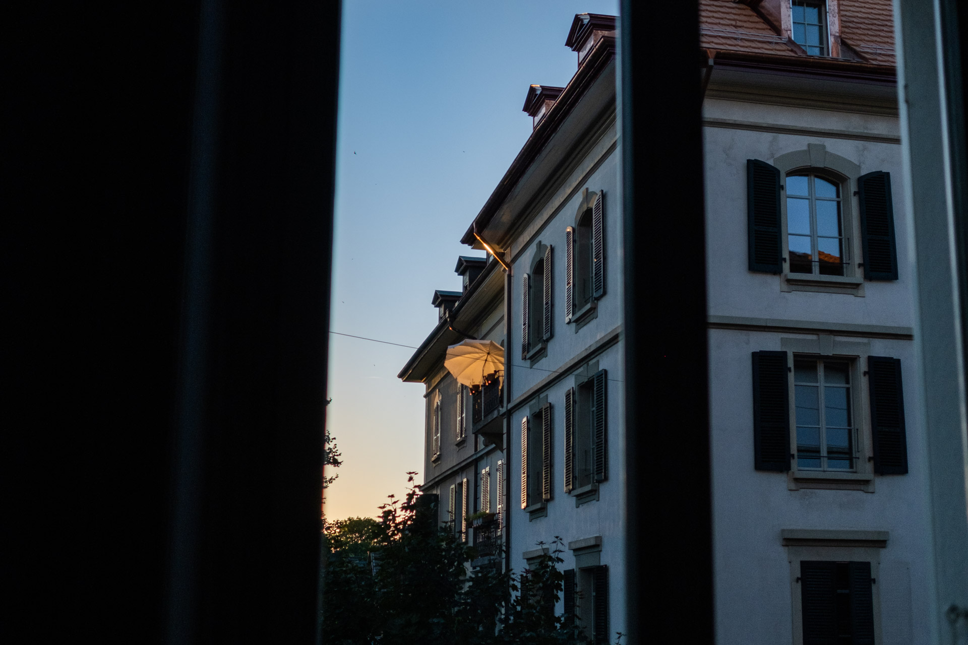 Blick aus dem Fenster auf ein benachbartes Altbau-Haus, bei dem auf einem Balkon ein oranger Sonnenschirm von der Abendsonne beleuchtet wird.