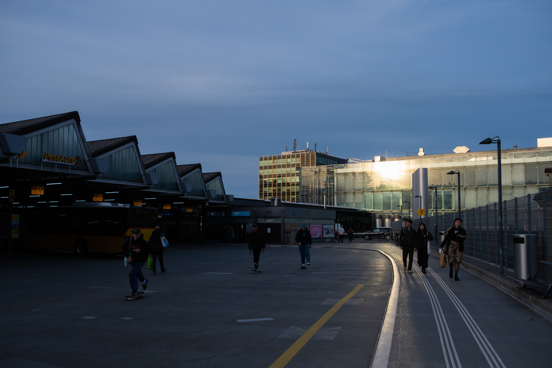 Abendstimmung am Bahnhof, mehrere junge Menschen verlassen die Haltestellen der Postautos. Im Bahnhofsgebäude spiegelt sich die Sonne im Glas.