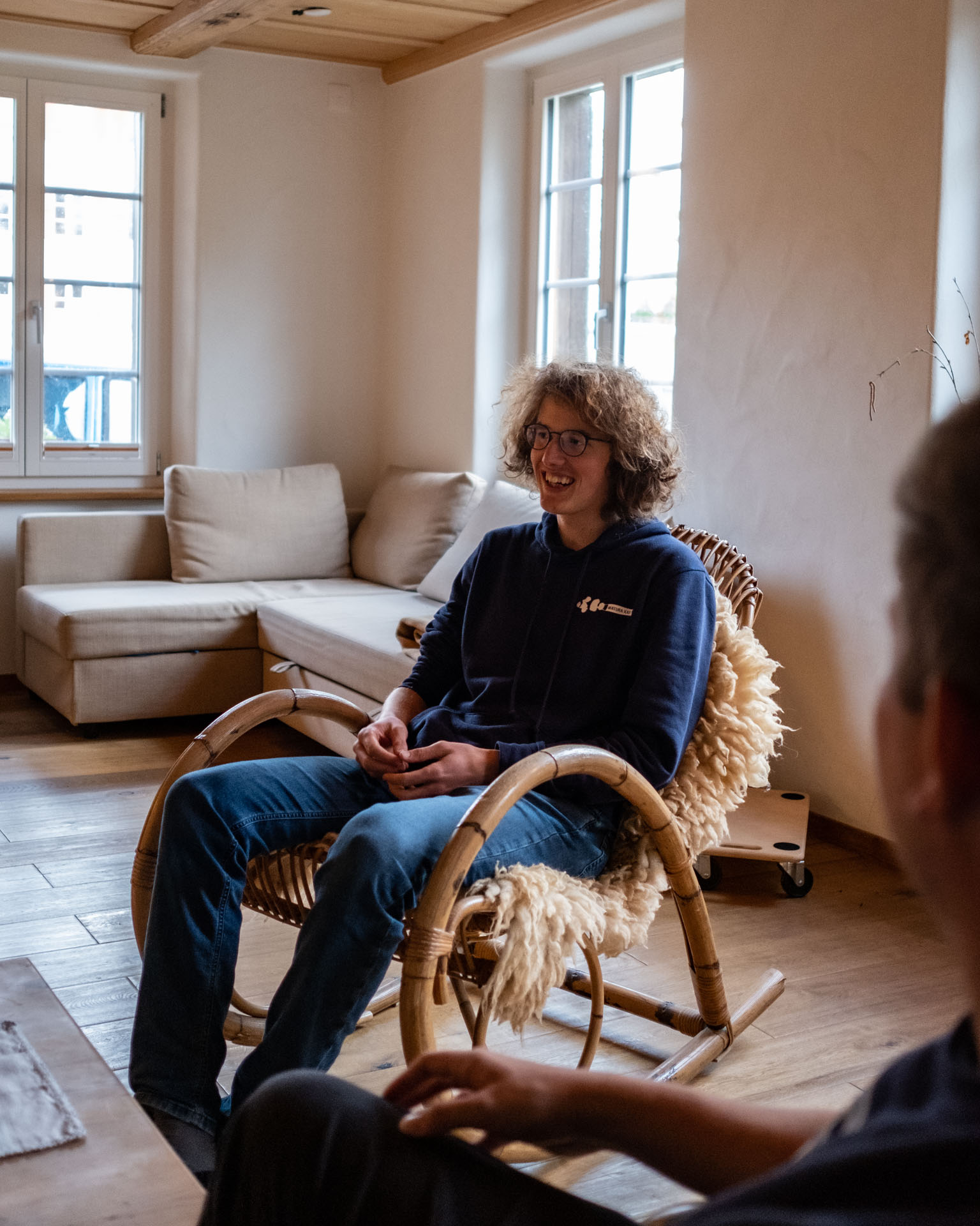 Ein junger Mann sitzt in einem geflochtenen Schaukelstuhl in einem gemütlich eingerichteten Zimmer.